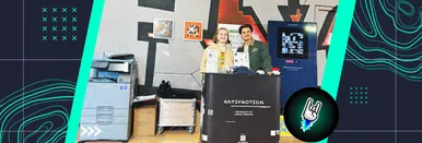 Roxy Josefus und Ares Avgerinos von 9elements stehen hinter dem Artifaction Stand bei der Millerntor Gallery 2025. Hinter ihnen sind Artifaction-Gemälde an der Wand befestigt, der Printer und der Generator sind ebenfalls zu sehen.