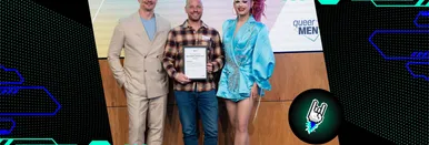 Preisträger Nils Binder hält die Urkunde des Queermentor Role Model Awards in der Hand und lächelt in die Kamera. Neben ihm stehen die Moderator*innen der Award-Show Rachel Intervention und Pavlo Stroblja.